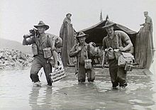 Black and white photo of three mean wearing military uniforms and carrying guns and bags wading through water in front of a boat