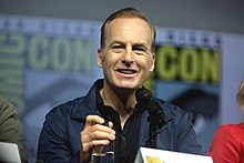 A man roughly in his mid-50s smiles straight ahead. In front of him, a microphone is visible, and in the background, the words "San Diego Comic Con" can be seen.