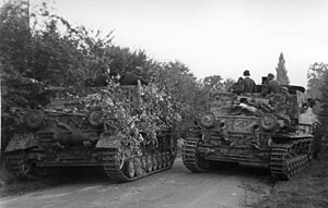 Two armoured vehicles one cowered in tree branches on a hedge lined road