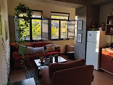 Room in Dialynas Hall with sofas, a coffee table, a refrigerator, and plants. There is light streaming through two open windows, a warm earth-toned color palette, and an exposed concrete column.