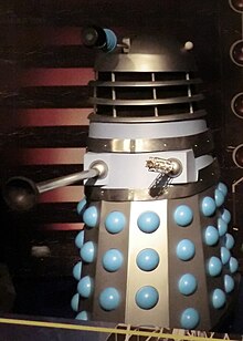 Un Dalek original, colorat în mare parte argintiu și gri, cu bile albastre care ies din fustă.