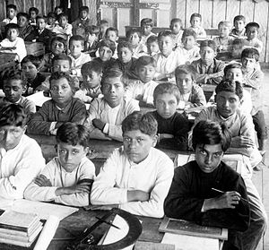 Black and white photo of dozens of students seated at desks in a classroom in Ecuador.