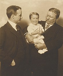 Theodore Roosevelt holds an infant, while Theodore Jr. looks on