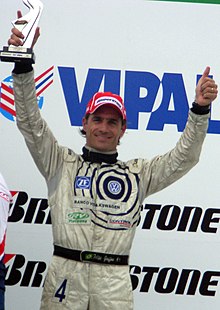 Fotografia de um homem, em plano americano, visto de frente, sorrindo, levantando um troféu.