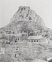 A black and white photograph showing a stone house alongside several other buildings, beneath a large stone mountain