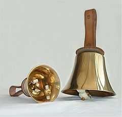 Zdjęcie przedstawia dwa dzwonki ręczne na białym tle. Dzwonki mają skórzane uchwyty. Instrumenty położone są obok siebie. Lewy z nich położony jest w pozycji poziomej, widziany od dołu. Drugi z nich położony jest w pozycji pionowej.