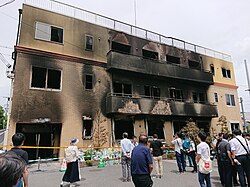 Na fotografii se nachází budova studia Kyoto Animation po žhářském útoku budovy, okna a dveře jsou zničené a fasáda spálená a zčernalá, a v její blízkosti se shromažďují lidé