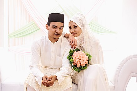 A Malay couple in traditional Malay clothings. The groom is wearing a Baju Melayu paired with songkok and samping, while the bride wears Baju Kurung with a tudong.