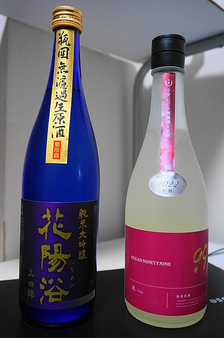 The blue sake bottle displays "Yamada Nishiki" (山田錦) and "Junmai Daiginjo" (純米大吟醸) on the bottom label and "Bingakoi muroka nama genshu" (瓶囲無濾過生原酒) and "requiring refrigeration" (要冷蔵) on the top label. The label on the pink sake bottle indicates Usunigori muroka nama genshu.