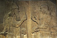 Ein Steinrelief zeigt Sargon II auf der linken Seite mit einer Krone und einem Stab gegenüber einem Mann auf der rechten Seite