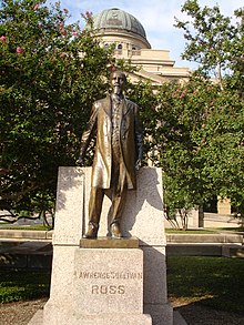 A large domed building overlooks a full-length statue of balding white male with a mustache and long goatee and wearing a knee-length coat. The pedestal is engraved "Lawrence Sullivan Ross".