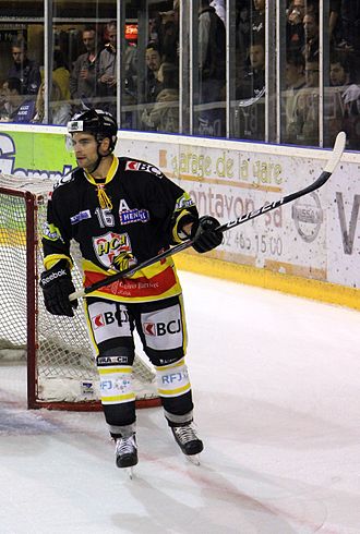 Giocatore di hockey di fronte, a piedi, vicino a una porta, il bastone tra le mani e alzato alla sua sinistra