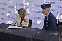 Seated woman signing agreement with man in military uniform seated to her right