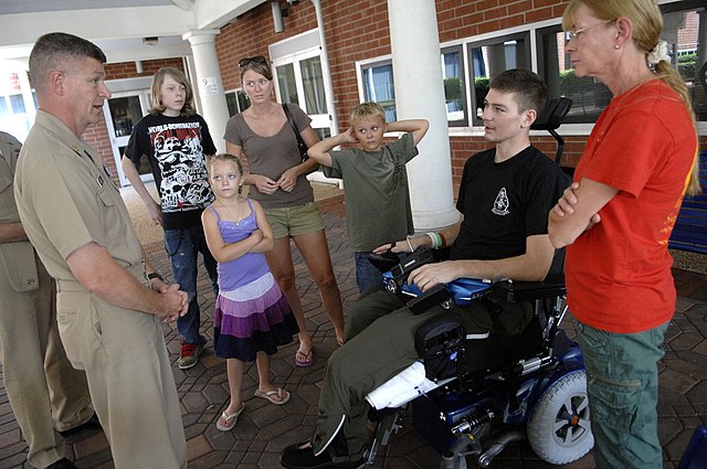 Color photograph of a man in uniform (left) talking to a man in an electric wheelchair (right) who is flanked by women and children.