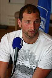 Farbfoto mit dem sitzenden Meynard, der ein weißes T-Shirt mit einer schwarzskizzierten aufgehenden Sonne trägt.