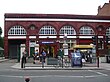 A red-bricked building with eight people standing in front of it and a blue sign reading "BELSIZE PARK STATION" in white letters all under a bright sky
