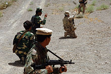 Photographie en couleur de cinq militaires portant deux uniformes différents avec une genou au sol pointant dans une direction.