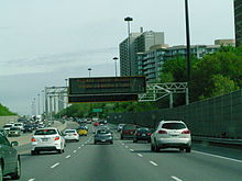 A large black digital message board is mounted onto a gantry over top of a set of freeway lanes.