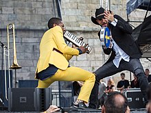 Jon Batiste in yellow clothes and Joe Saylor in black clothes and cowboy hat, playing a tambourine