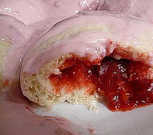 Zdjęcie pyzy drożdżowej z nadzieniem truskawkowym. Kluska polana jest różowym sosem jogurtowym, jest w połowie zjedzona, z jej wnętrza wypada rozgotowana truskawka. Nadzienie ma intensywny czerwony kolor. W tle inne pyzy polane sosem.