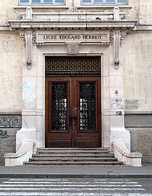 Entrée du lycée Édouard-Herriot. Une porte massive en bois, à double battant, au-dessus de laquelle est gravé le nom du lycée, donne sur quelques marches puis le trottoir.