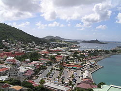 A village on the coast of an island. Small buildings are located throughout the island, with mountains in the background and the ocean on the right. Among the buildings in the foreground is a parking lot adjacent to a marina. A peninsula stretches out into the ocean and boats are on the ocean in the background.