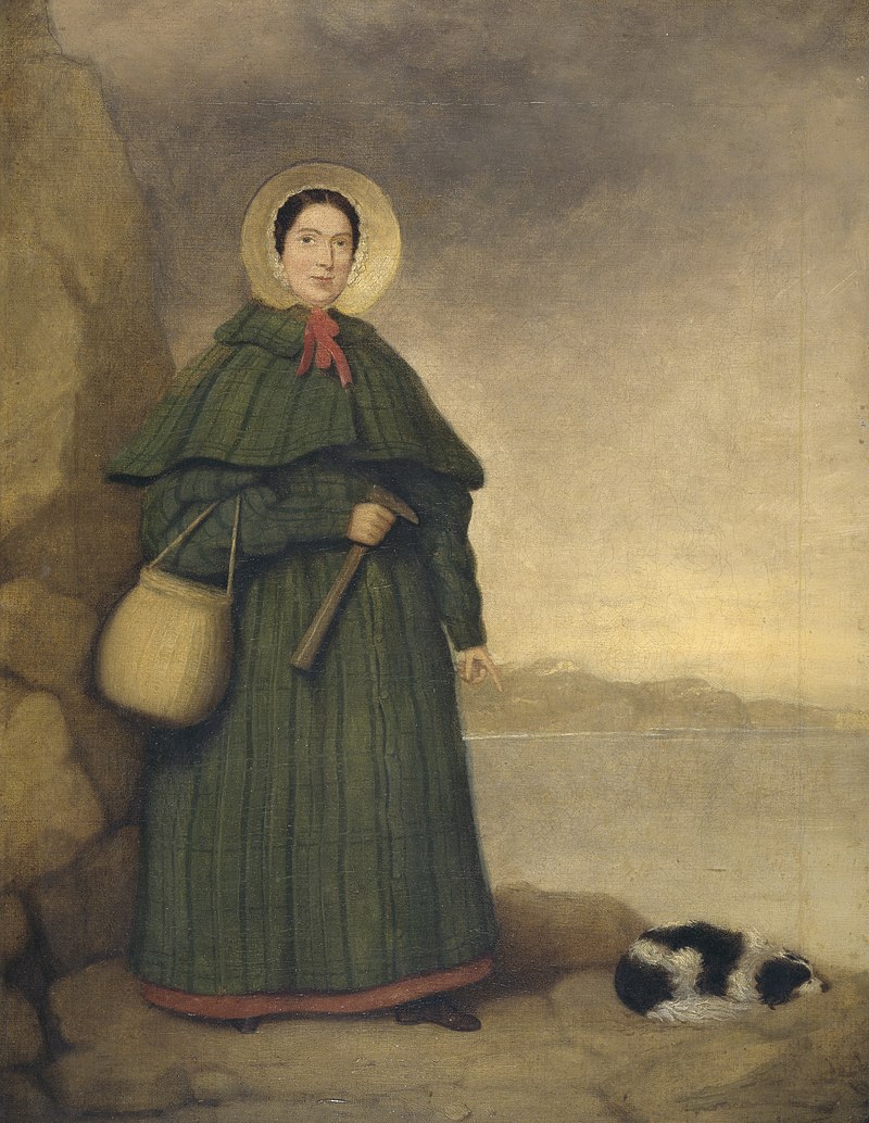 Portrait of a woman in bonnet and long dress holding rock hammer, pointing at fossil next to a spaniel dog lying on ground.