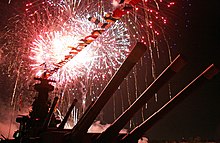 Fogos de artifício vermelhos e amarelos enchem o céu, visto do convés do navio.  Os canhões principais aparecem diretamente acima.