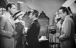 Black-and-white film screenshot of several people in a nightclub. A man on the far left is wearing a suit and has a woman standing next to him wearing a hat and dress. A man at the center is looking at the man on the left. A man on the far right is wearing a suit and looking to the other people.