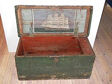 An old looking chest with weathered green paint sits open. There is a decorative painting of a large sailing ship on the inside of the lid with "Auguste Victoria" written under it.