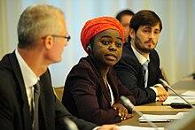 Photograph of a Black woman wearing a dark suit and with a red head wrap speaking into a microphone on a panel. On either side of her is a white man in a suit.