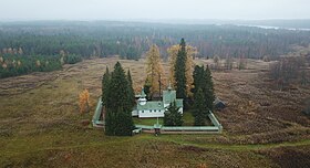 Vue aérienne d'une église en bois entourée d'une clôture carrée, avec autour une prairie puis des étendues de taïga.