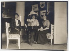 Three white women seated at a table indoors; two are wearing large hats