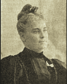 B&W portrait photo of a woman with her blonde hair in an updo, wearing a dark blouse with a white ribbon pinned on it.