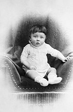 Photo en noir et blanc d’Adolf Hitler prise peu après sa naissance. Sur un fond blanc, dans un médaillon au fond sombre, au centre de la photo, un bébé, en layette blanche, est assis sur un siège en velours foncé. Les yeux grands ouverts, il fixe l’appareil photo avec curiosité.