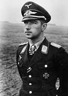 Black-and-white photograph showing the face and upper body of a young man in uniform, his hands behind his back, standing in a featureless landscape. His cap and the front right of his jacket bear eagle-and-swastika emblems; the front left of his jacket and the front of his shirt collar bear Iron Cross decorations, black with light outline. He is shown in semi-profile, gazing at a point in the distance to the left of the camera, his facial expression confident.