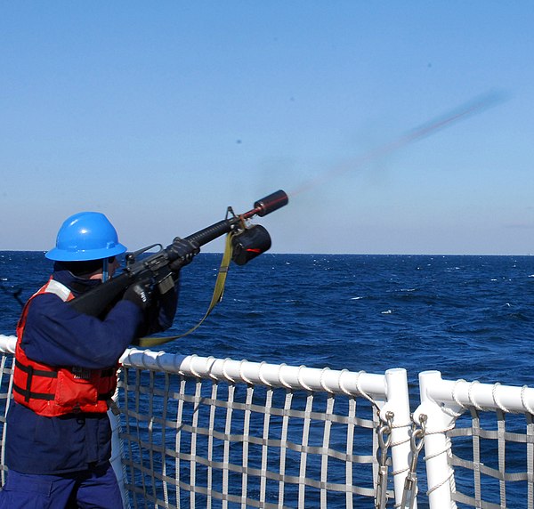 A USCG sailor uses a specially adapted firearm to fire a messenger line to another vessel.