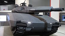Makieta polskiego czołgu lekkiego PL-01 ustawiona w hali wystawowej. Pojazd ukazany z bliska, od frontu, sprzed prawej gąsienicy.