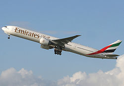 Plejparte blanka Boeing 777, kun kelkaj ruĝaj, verdaj kaj nigraj markaĵoj, de Emirates Airlines, en flugo, alfrontante maldekstron.