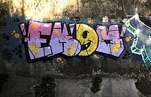 picture of a graffiti piece done using the letters EKOM. the E, K, and M and purple and pink. The O is a yellow smiley face.