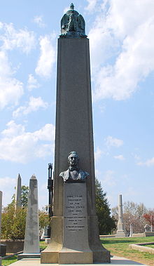 Fotografia de um obelisco cinza localizado em um cemitério atrás de um busto de Tyler.