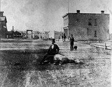 A picture of a man in a suit and a top hat sitting on the body of a dead horse in an old-looking stone road