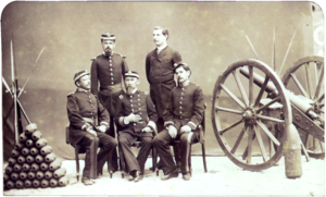 A photograph depicting a group of 5 uniformed men posed between a pyramid of artillery shells on the left and a wheeled field artillery piece on the right