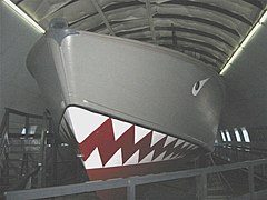 An open boat sits inside a museum building. The area above the waterline is gray, that below the waterline is red with shark teeth. An eye is also painted on the gray part of the ship, making it look a little like a shark. A walkway goes around the boat.