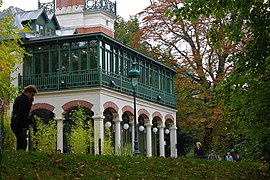 Farvefoto af en bygning med søjler og arkader i polykrome mursten, der understøtter en loggia og en grøn træhave.