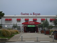 Casino de Royan'ın iki seviyede yatay beyaz bir binayı gösteren fotoğrafı.  Girişi gösteren gölgelik, üst kattaki panjurlar ve düz çatıdaki tabela kırmızıdır.