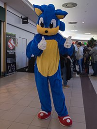 An individual dressed up as a cartoon blue hedgehog with a tan-colored muzzle and stomach, white gloves, and red shoes.