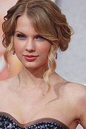 Taylor Swift, wearing a strapless dress, looks directly at the camera. Her hair is tied back, with a few curly tendrils loose