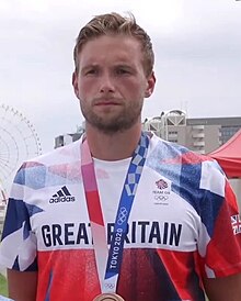 Thomas Ford trägt das britische Olympiatrikot. Um den Hals trägt er ein blaues Band, an dem seine Bronzemedaille befestigt ist, die aber nur zum Teil im Bild ist.