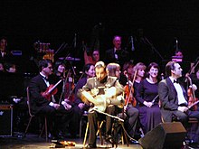 A slightly blurry image of an orchestra, with Rony Barrak pictured in the centre playing a goblet drum